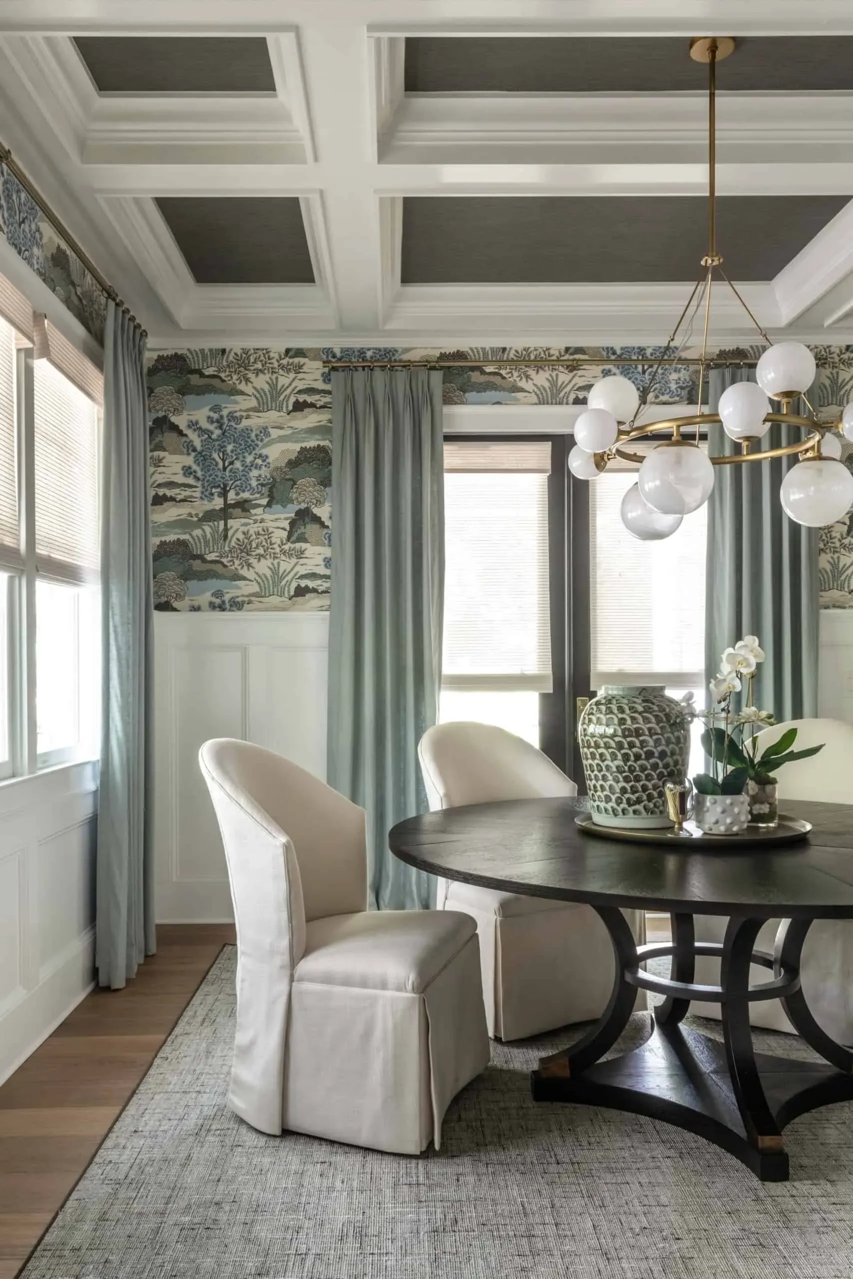 Soft blue-green linen drapes with woven shades, hung from a brass rod across multiple windows in a dining room with chinoiserie wallpaper and a coffered ceiling.